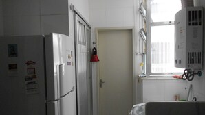 A narrow kitchen with white cabinets, a refrigerator, and a window with blinds. - Gohouse - Siqueira 703 (Rio de Janeiro)