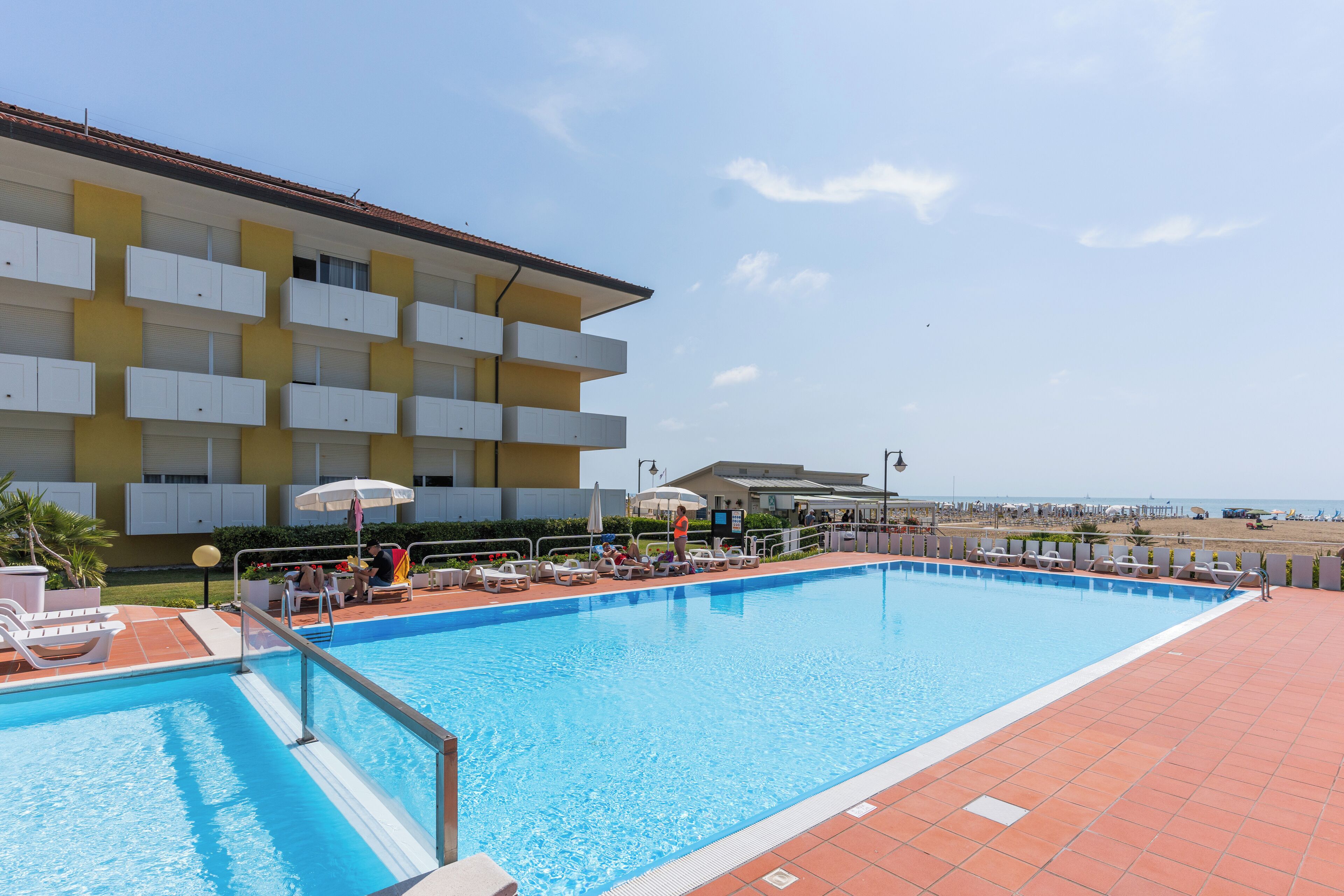 A swimming pool with lounge chairs and umbrellas, a building with balconies, and a beach in the background.