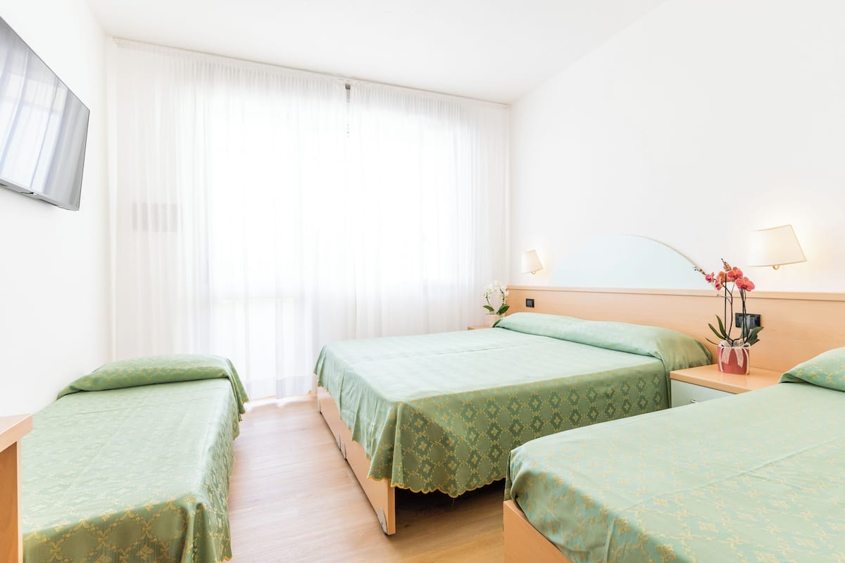A hotel room with two single beds, a bedside table with a lamp, and a large window with sheer curtains.
