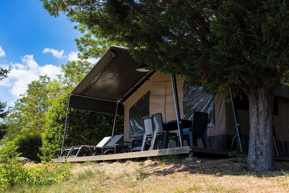 Een tentaccommodatie met een overdekt terras, lounge stoelen en een boom in de buurt.