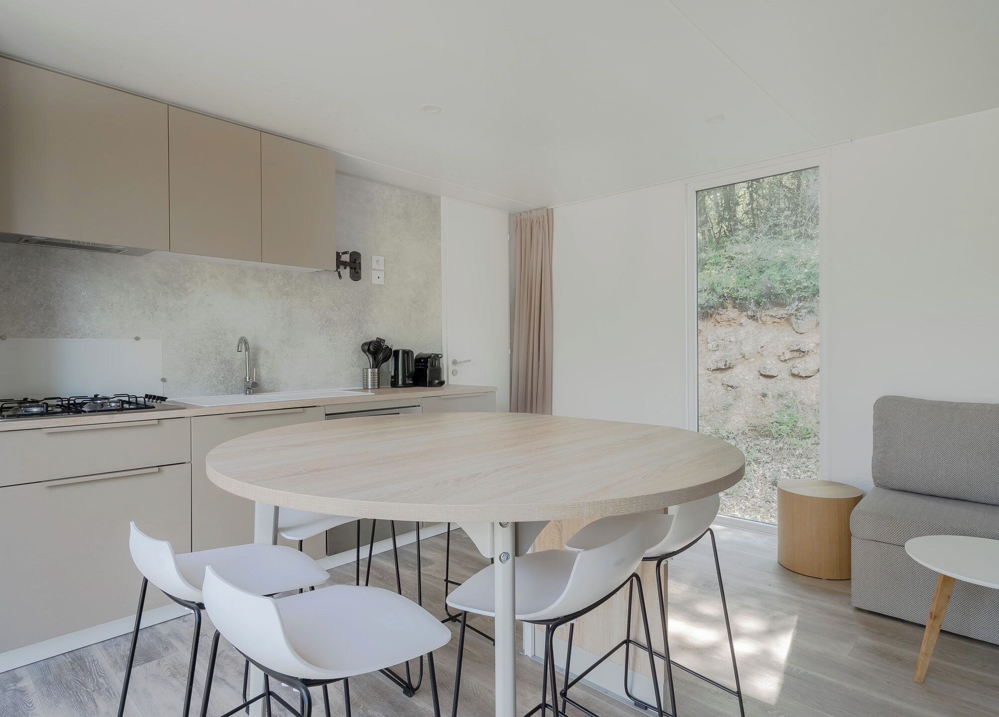 Cozinha moderna com mesa de jantar redonda e cadeiras brancas, um sofá e uma janela grande com vista para áreas verdes.