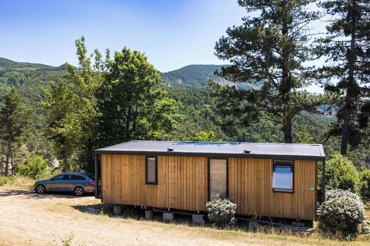 Een klein houten huisje met een metalen dak, gelegen in een bebost gebied met bergen op de achtergrond.