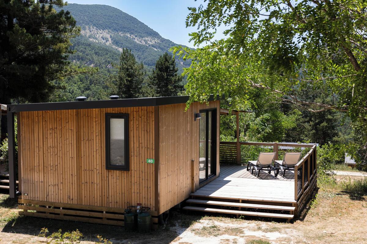 Een houten chalet met een terras, twee stoelen en uitzicht op een bebost berglandschap.