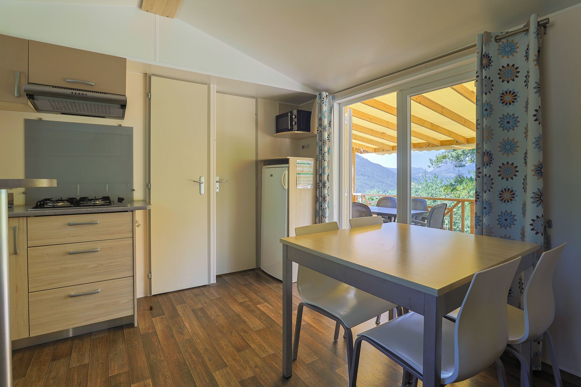 Eine kompakte Küche mit Gasherd, Mikrowelle und Kühlschrank. Es gibt einen Essbereich mit Tisch und Stühlen, sowie ein Fenster mit Blick auf Grünflächen und Berge.