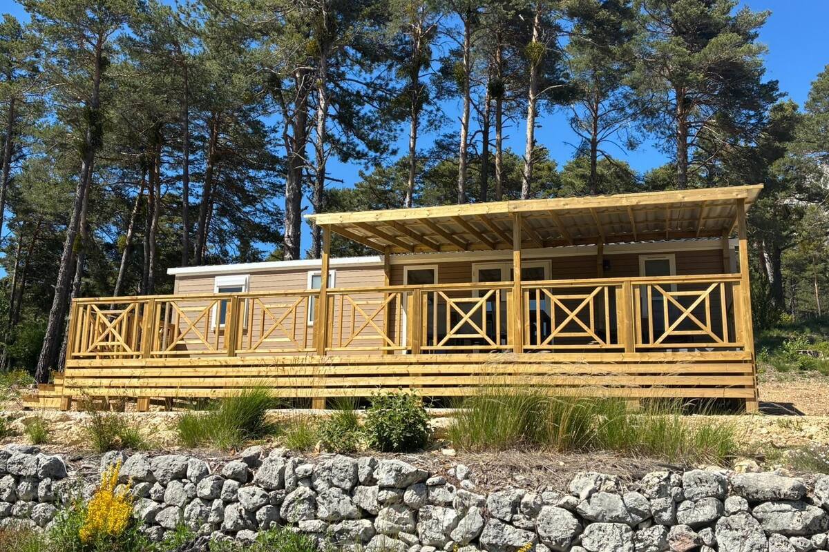 Een houten hutje met een terras, omgeven door een stenen muur en pijnbomen.