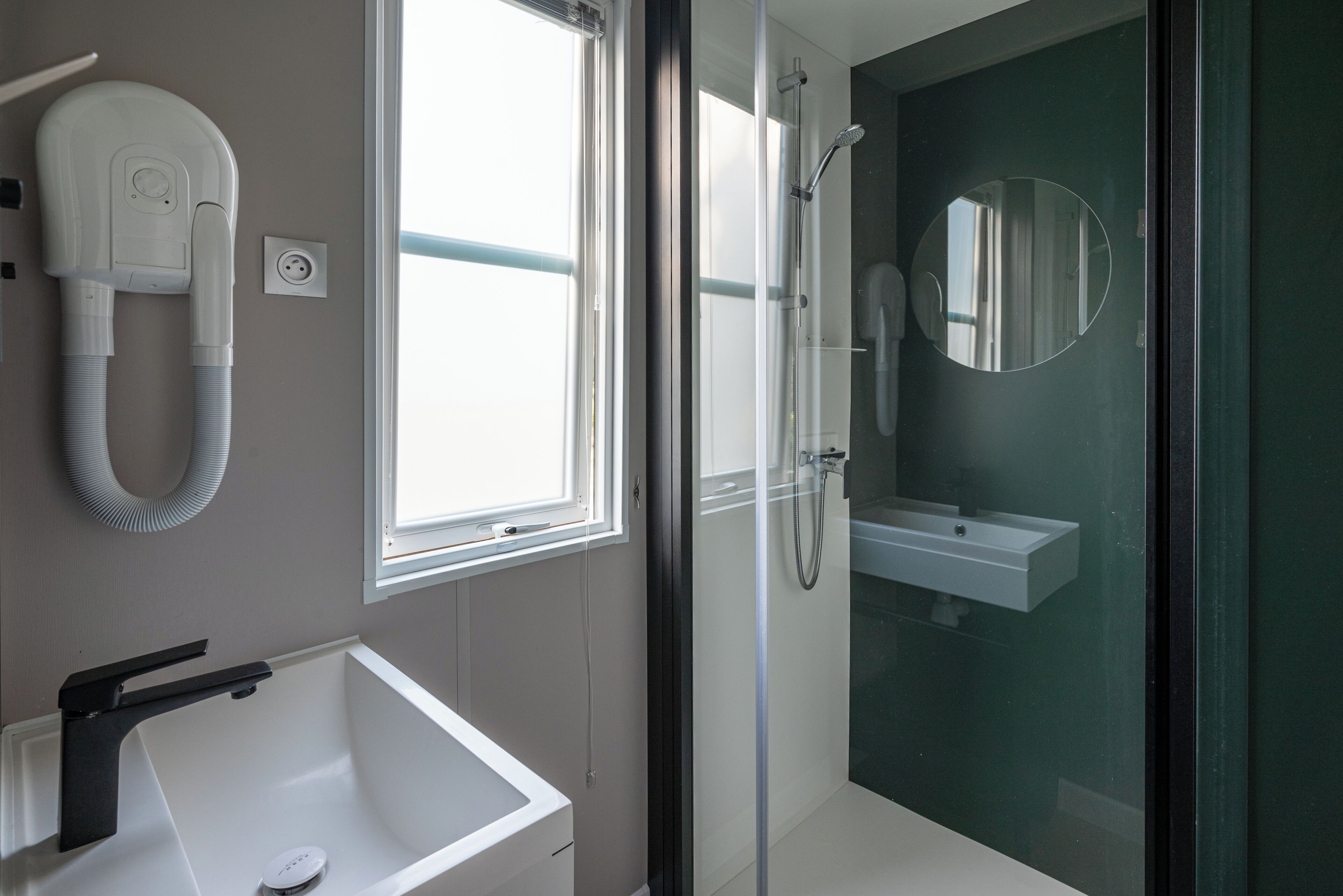 A modern bathroom with a white sink, a hairdryer, a glass shower enclosure, and a mirror.