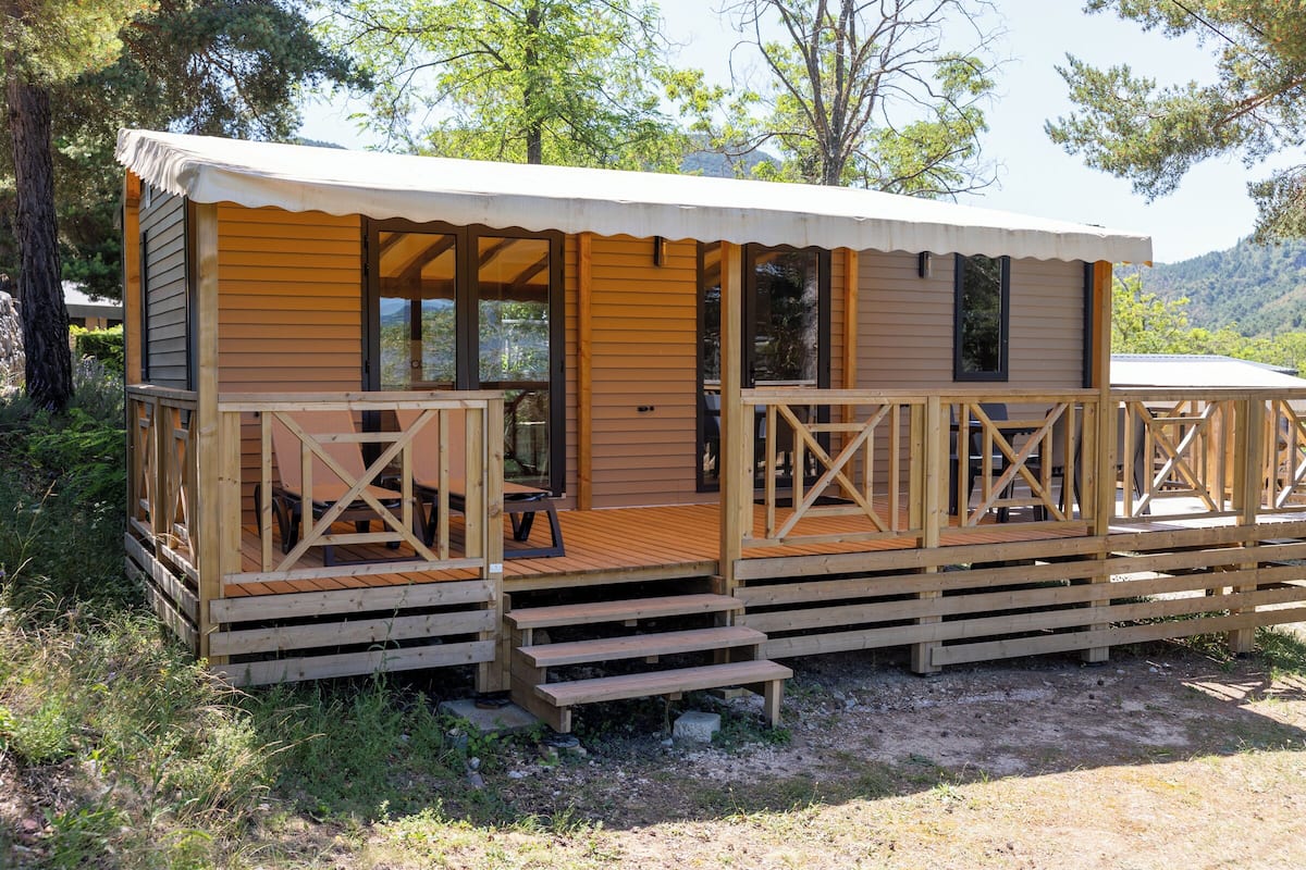 Een houten hut met een overdekte veranda en een terras, omringd door bomen en een glooiend landschap.