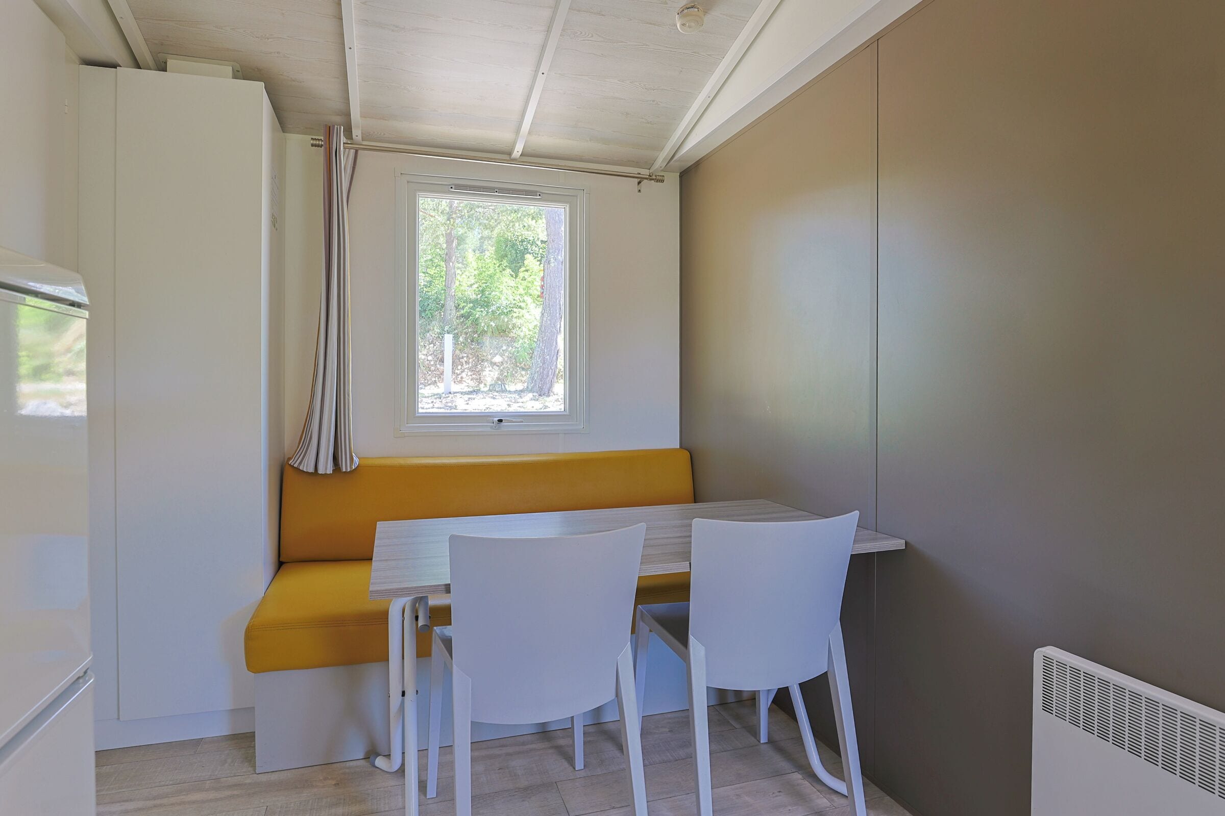 Een compacte keuken met een inbouwbank en witte stoelen, een raam met gordijnen en een witte koelkast.