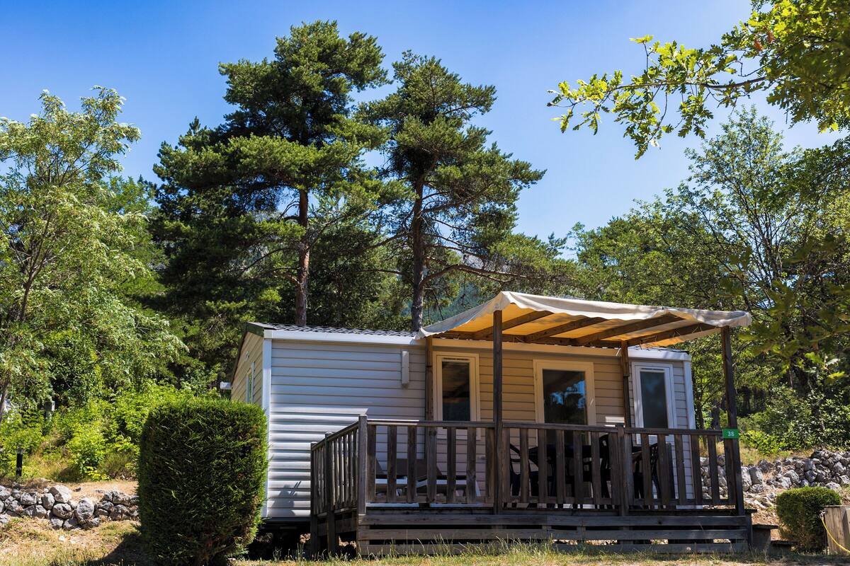Een klein vakantiehuisje met een houten terras en een overdekte veranda, omringd door bomen en een stenen muur.