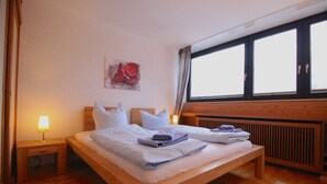A hotel room with a wooden bed, a nightstand with a lamp, a radiator, and a window with curtains. - Wettersteinblick (Garmisch-Partenkirchen)
