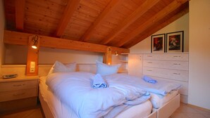 Une chambre avec un plafond en bois, un lit recouvert de linge de lit blanc, une table de chevet, une commode et deux tableaux encadrés accrochés au mur.
