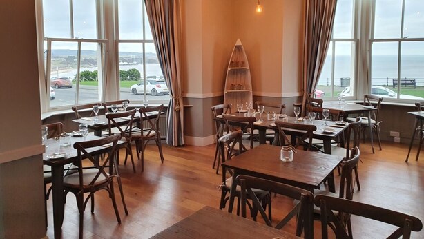 A dining area with wooden tables and chairs, a central wooden shelf, and large windows with a view of the sea. - Delmont Hotel (Scarborough)