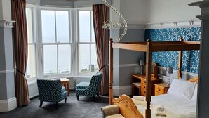 A four-poster bed with a canopy, a sitting area with a patterned armchair, a small table, and a view of the sea through large windows. - Delmont Hotel (Scarborough)