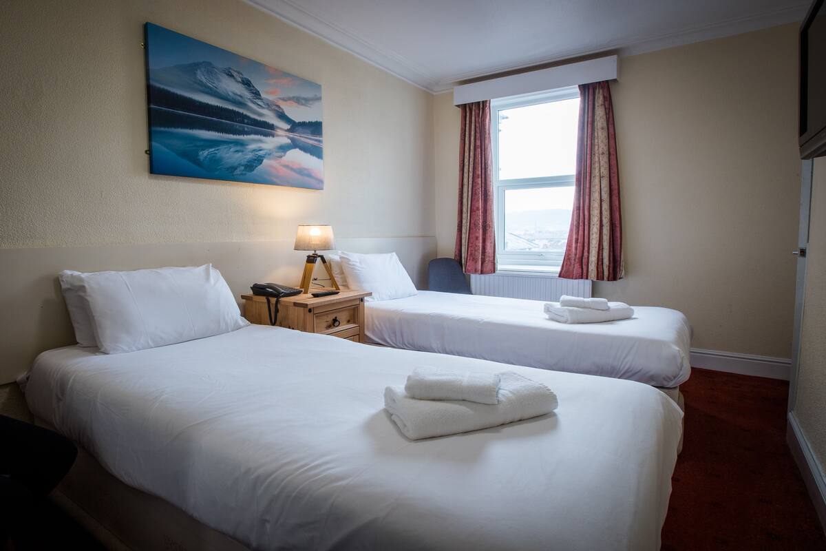 Hotellihuone, jossa on kaksi sänkyä, seinällä taulu, ikkunassa verhot ja yöpöydällä lamppu.