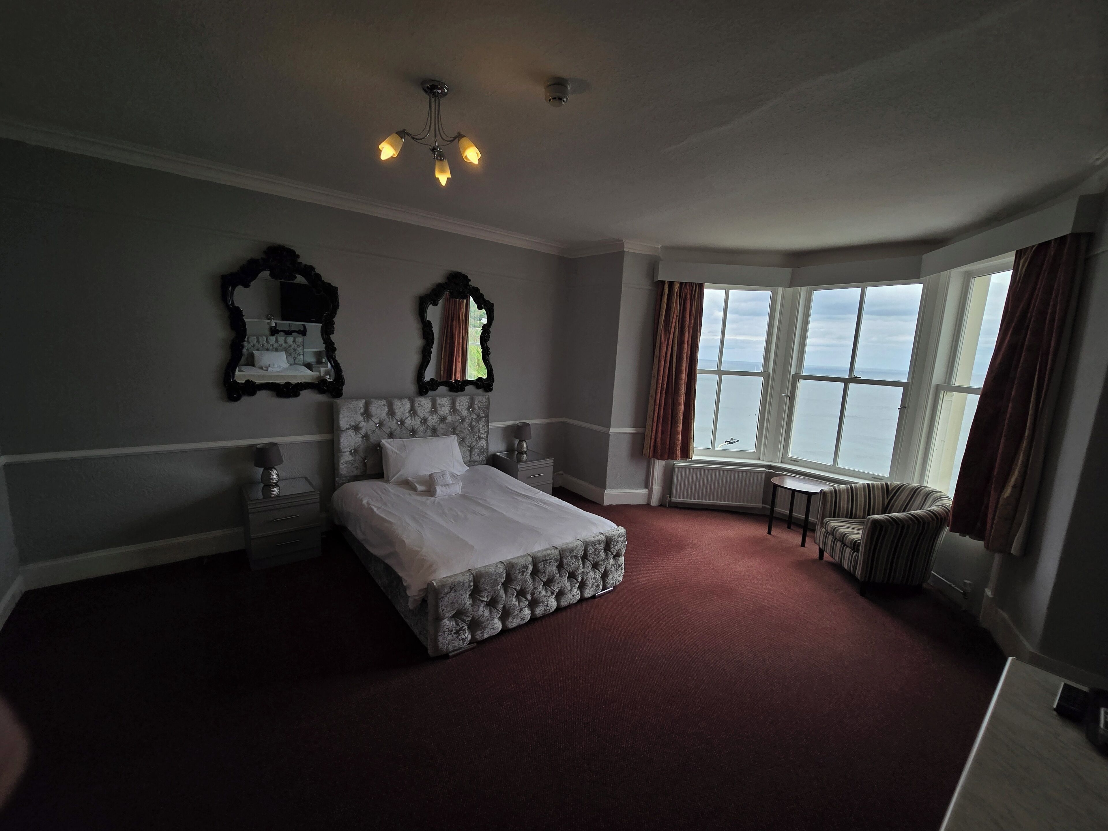 A hotel room with a large bed, a small table, a chair, and a view of the sea through a large window.