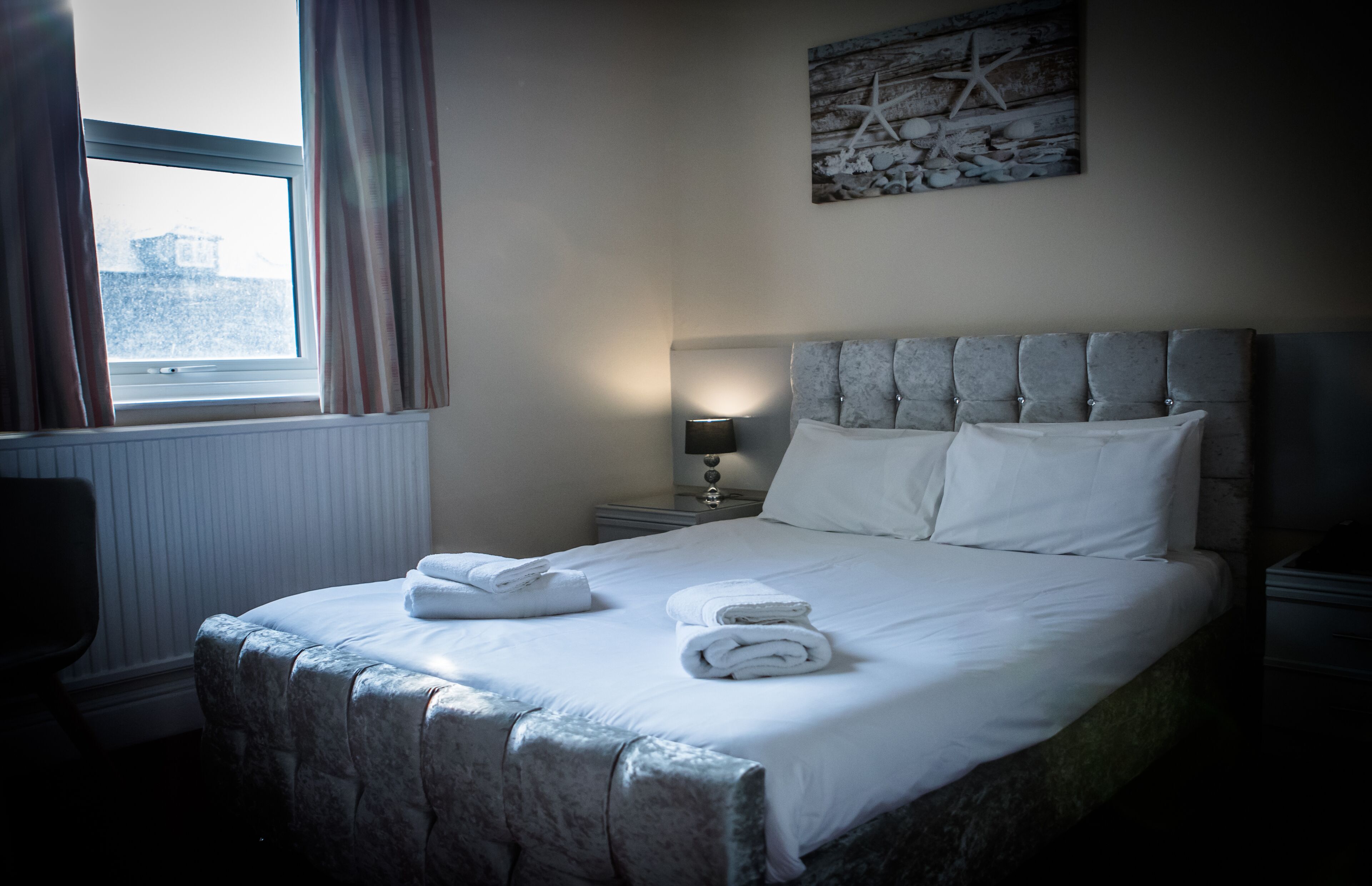 A neatly made bed with towels, a headboard, a nightstand with a lamp, a window with curtains, and a wall decoration.