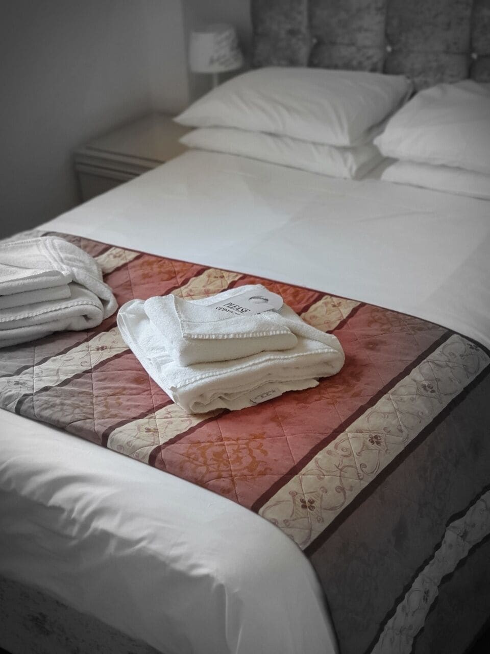 A neatly made bed with a quilted bedspread, a stack of folded towels, and a pillow on a nightstand.
