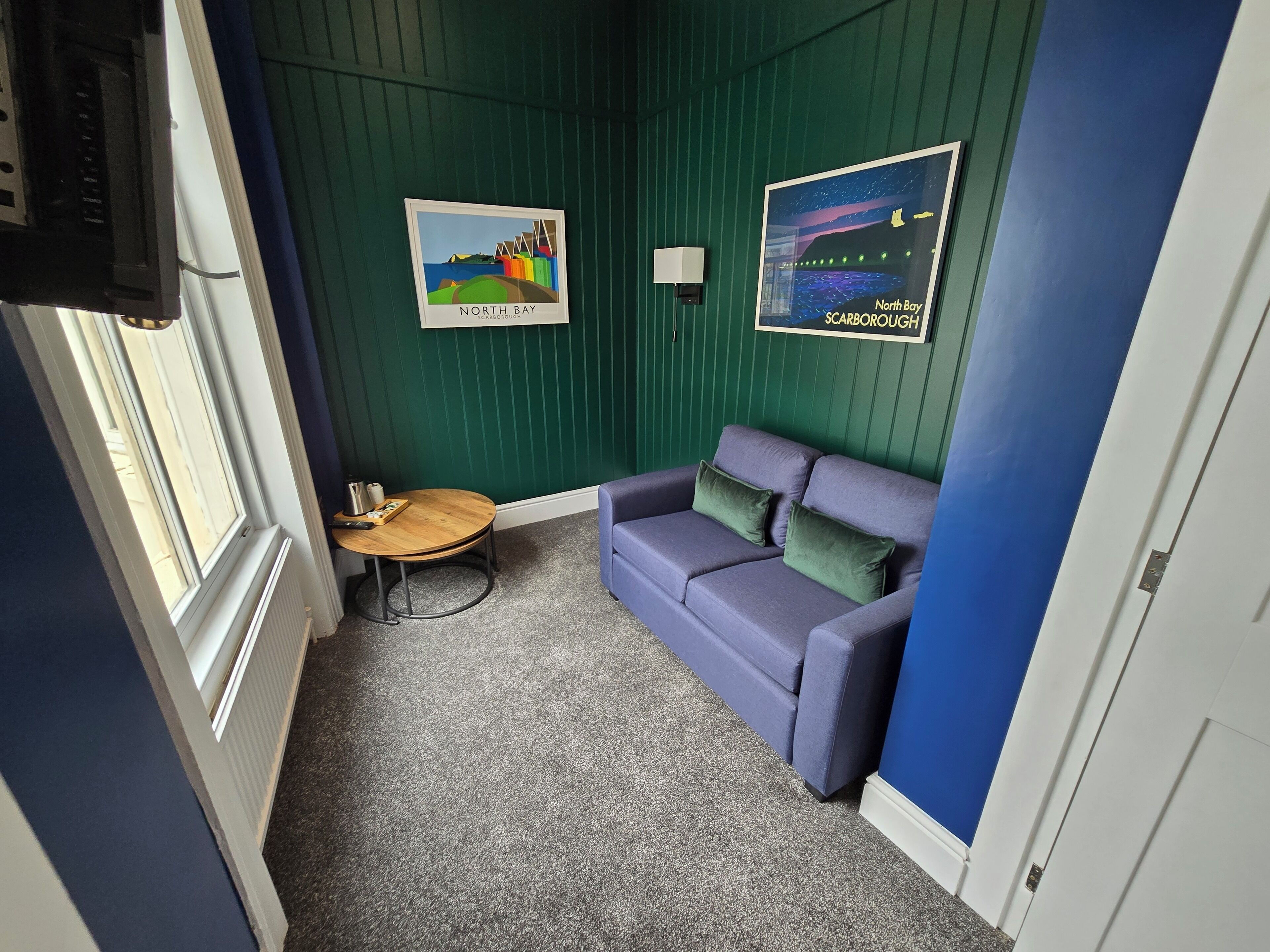 Una habitación pequeña con pared azul, alfombra gris, sofá púrpura, una mesita redonda y dos cuadros enmarcados.