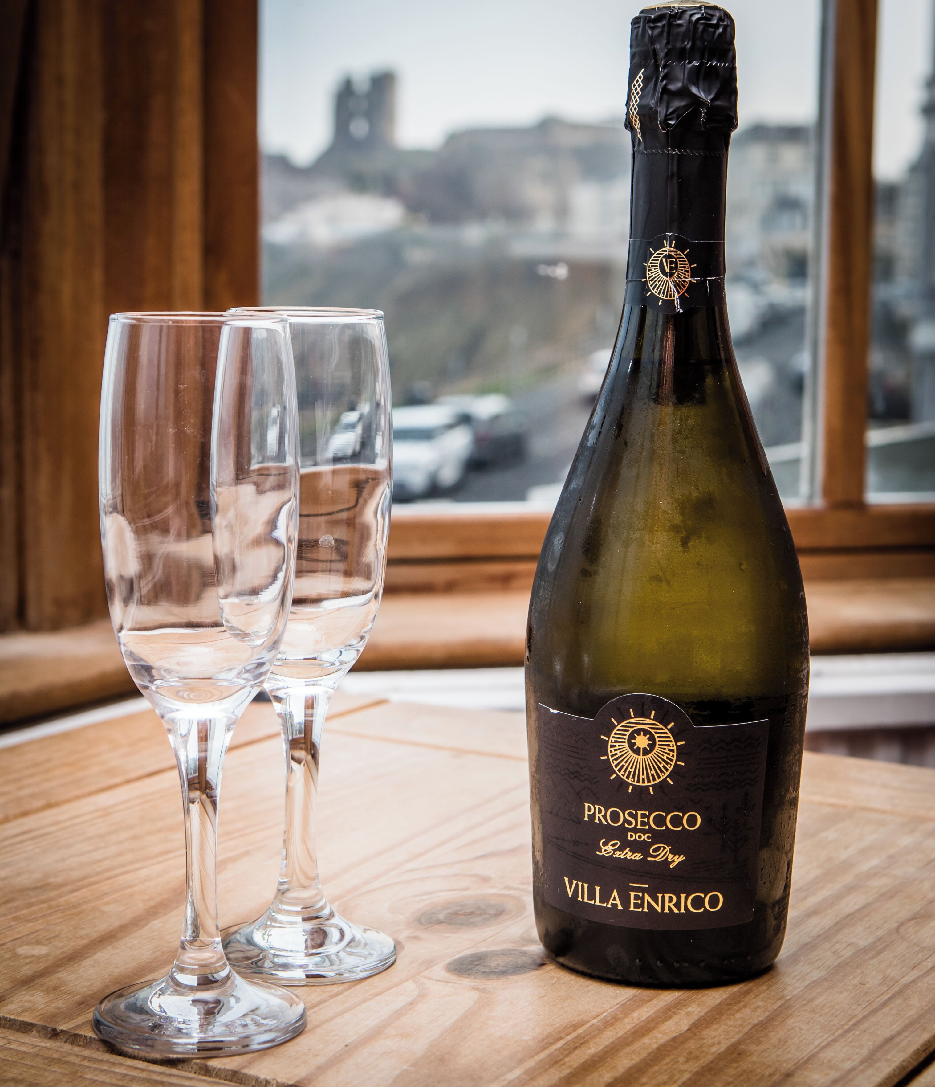 A bottle of Prosecco Doc Extra Dry Villanrico wine and two empty wine glasses on a wooden table by a window with a view of a cityscape.