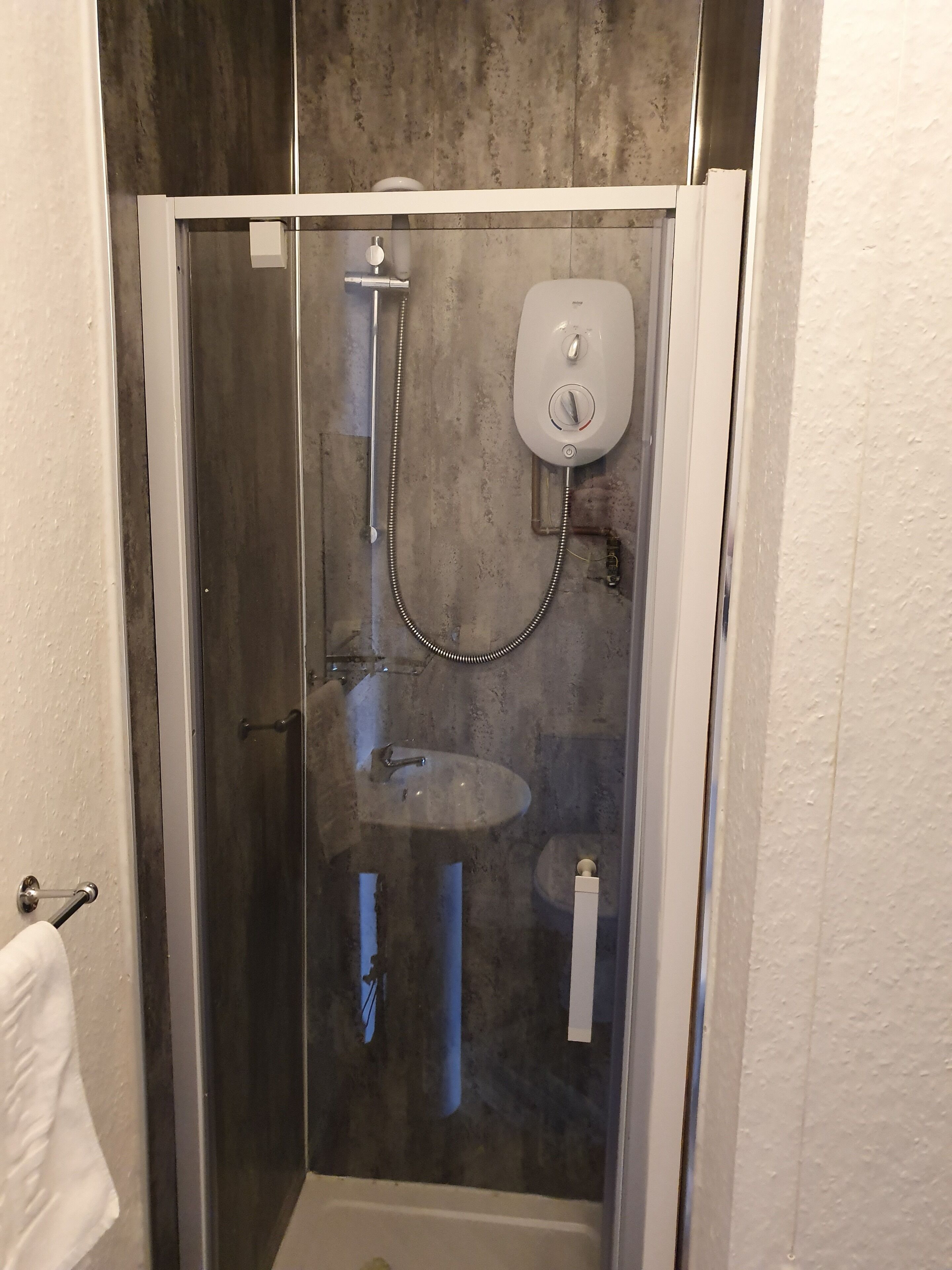 A glass-enclosed shower with a handheld showerhead, a wall-mounted soap dispenser, and a small sink.
