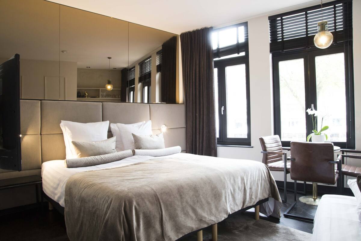 A modern hotel room with a large bed, a seating area with a table and chairs, a large window with blinds, and a mirror above the bed.