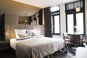 A modern hotel room with a large bed, a seating area with a table and chairs, a large window with blinds, and a mirror above the bed.