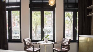 A hotel room with a bed, a round table with two chairs, a potted plant, and large windows with a view of buildings and trees. - House of Amstel (Amsterdam)