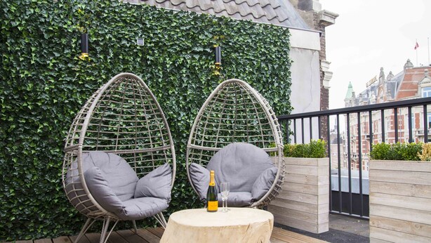 A wooden deck with two wicker chairs and a round table, set against a backdrop of a green ivy wall and a cityscape. - House of Amstel (Amsterdam)