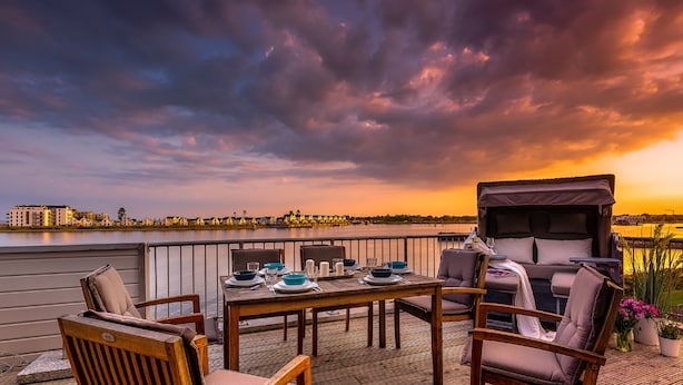 A terrace with a table set for dining, overlooking a body of water and a city skyline at sunset. - Viktoria's Baltic Lounge - First row on the water, sauna, fireplace, balcony (Olpenitz)