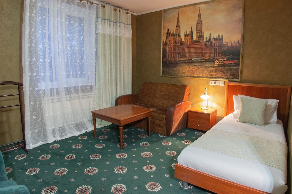 Habitación de hotel con cama, escritorio, silla, mesita de noche con lámpara, un cuadro de un paisaje urbano y alfombra estampada.
