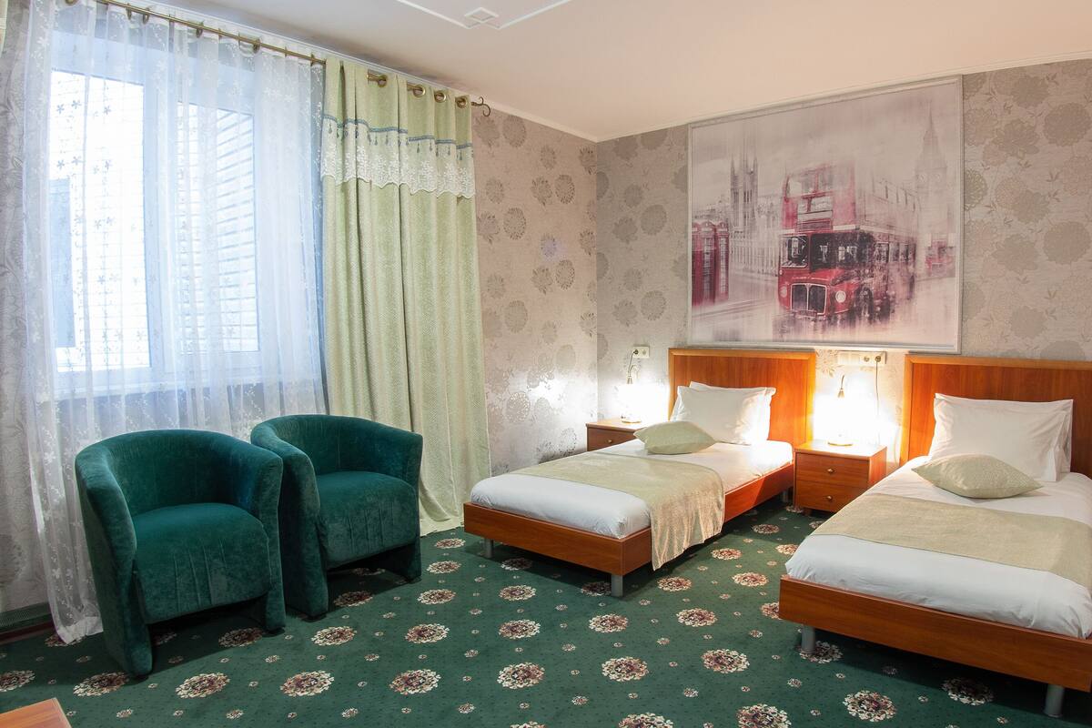 Una habitación de hotel con dos camas individuales, un par de sillones verdes, una alfombra de estampado floral grande y un cuadro con un paisaje urbano en la pared.