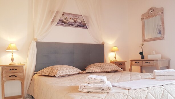A neatly made bed with a quilted duvet, two pillows, and folded towels. There are bedside tables with lamps, a mirror, and a small plant on the nightstand. - Emmanouil Airport Beach Apartment (Hersonissos)
