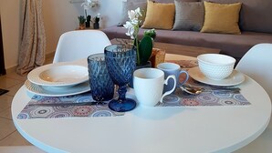 A dining area with a round table set for two, featuring plates, glasses, and a bowl. A sofa and a mirror are in the background. - Emmanouil Airport Beach Apartment (Hersonissos)