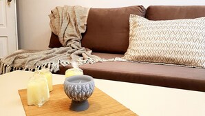A living room with a brown sofa, patterned cushions, a mirror on the wall, and a table with candles and a decorative bowl. - Emmanouil Airport Beach Apartment (Hersonissos)
