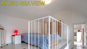 Ein Schlafzimmer mit Bett, Nachttisch in Weiß, roter Lampe und Meerblick durch ein Fenster.