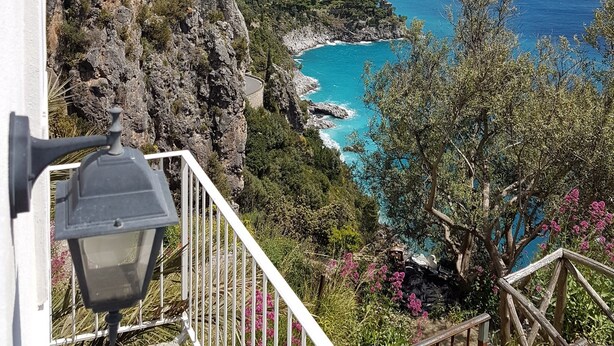 Blick auf die Küste mit einer Treppe, die zu einem Balkon mit Blick auf türkisfarbenes Wasser und eine gebirgige Landschaft führt.