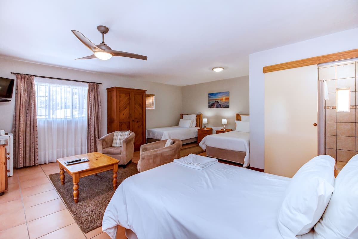 A hotel room with a bed, two bedside tables, a chair, a small table, a ceiling fan, a TV, and a window with curtains.