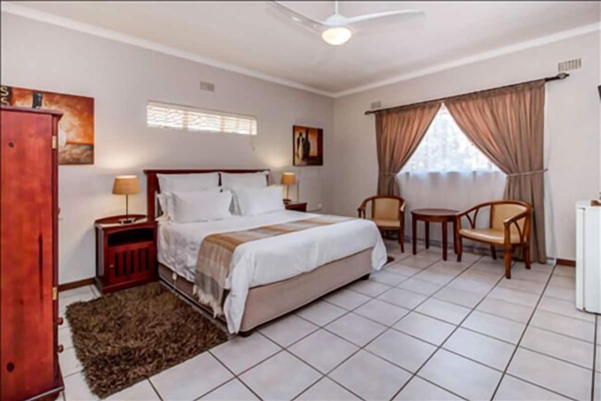 A bedroom with a bed, two bedside tables, a chair, a small table, a radiator, and a window with curtains.