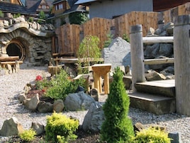 Un espacio al aire libre rústico con un arco de piedra, cerca de madera y varios elementos de jardín como rocas, plantas y un árbol pequeño.
