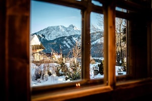Vista desde una ventana a un paisaje montañoso con picos nevados y un edificio rústico con tejado de paja.