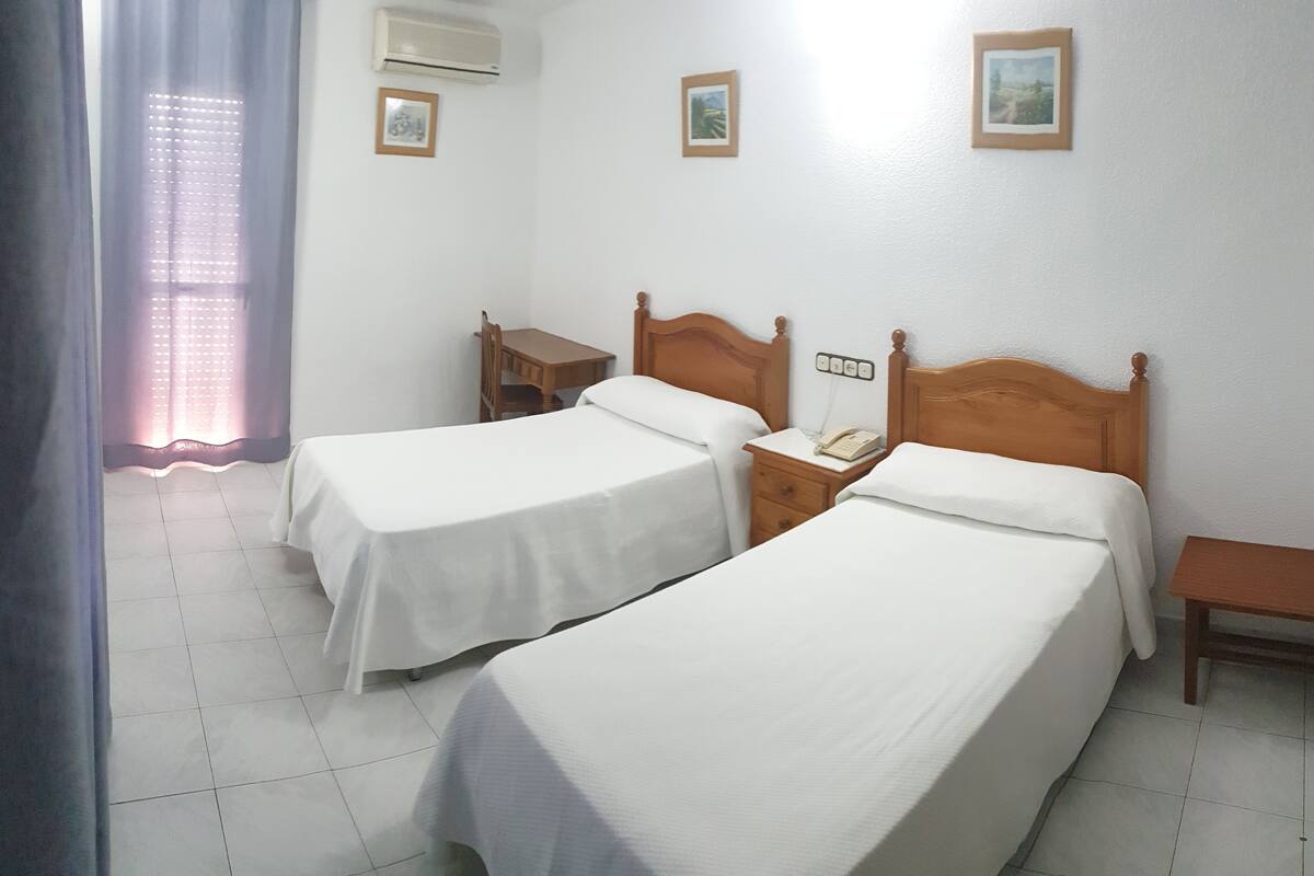 Een hotelkamer met twee eenpersoonsbedden, een nachtkastje van hout met een telefoon, een kleine tafel en een raam met gordijnen.