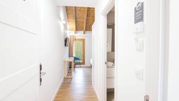 Un pasillo estrecho con suelo de madera y paredes blancas, que conduce a una habitación con techo de madera y ventana.