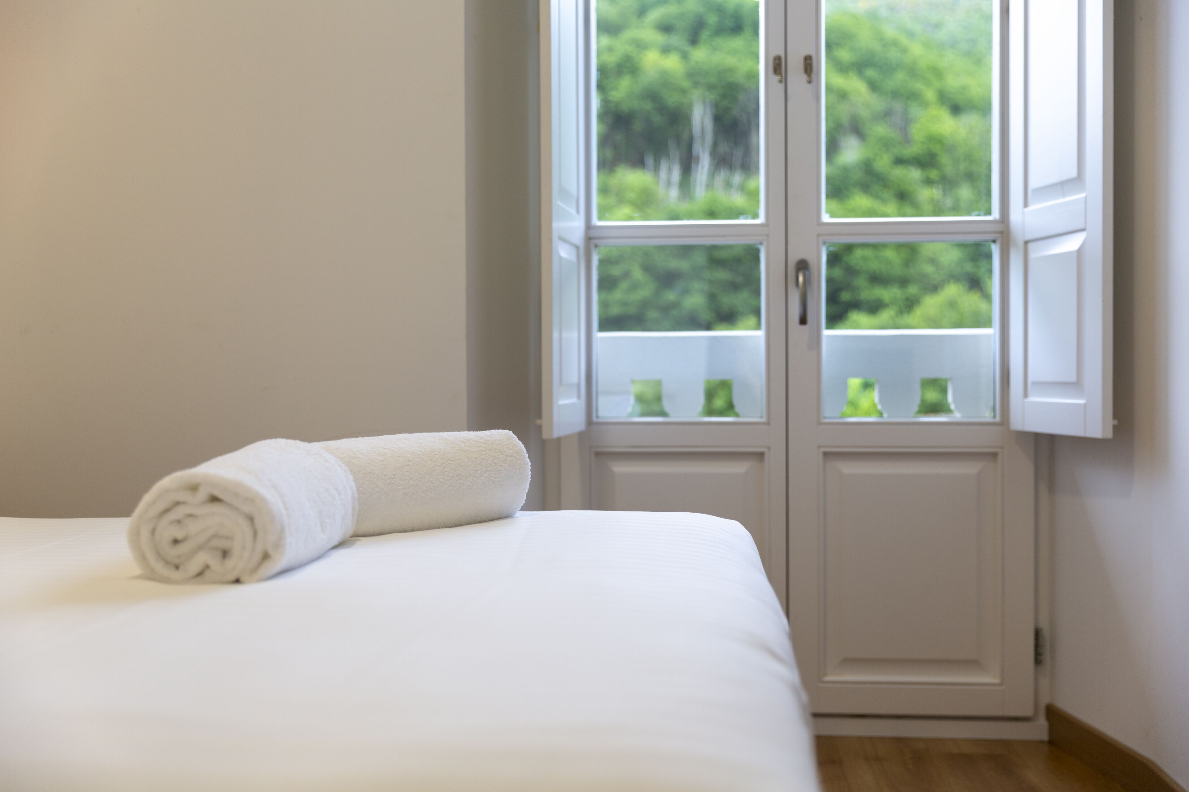 Una cama bien hecha con una toalla blanca enrollada, una puerta blanca que ofrece vistas al verdor exterior y una pared blanca.
