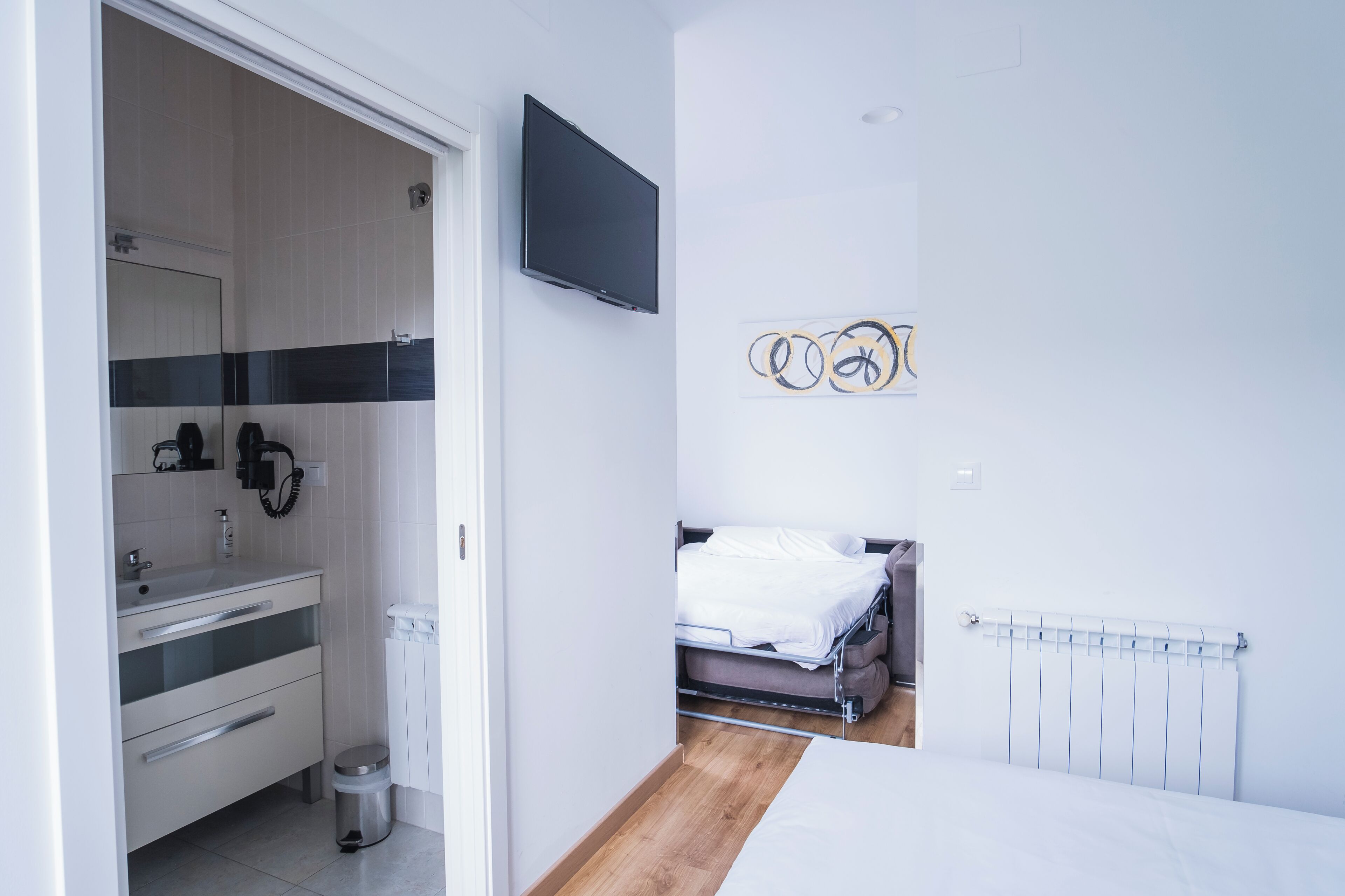 Un dormitorio moderno con cama, televisor montado en la pared, radiador y baño con lavabo y espejo.