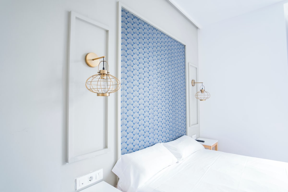 Un dormitorio con una cama blanca, un cabecero azul con estampado y dos lámparas fijadas a la pared.