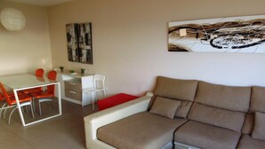 A modern living room with a sofa, a dining table with chairs, a small white desk, and a painting on the wall.
