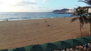 A beach with umbrellas and tables, a view of the sea, and buildings in the background.