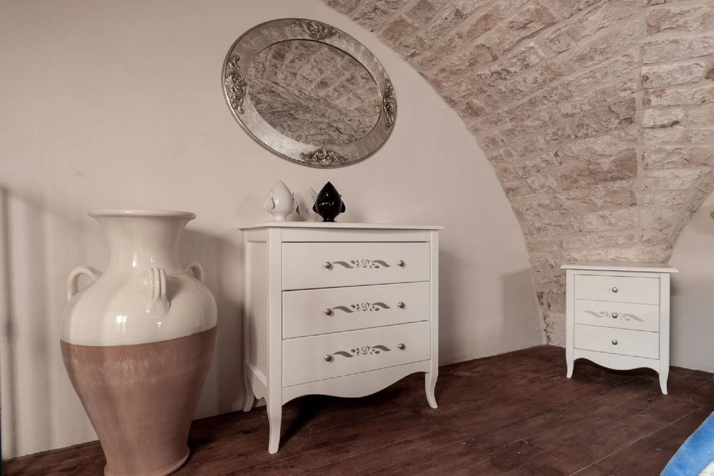 Quarto com cômoda branca, mesa de cabeceira pequena e um vaso grande em assoalho de madeira.