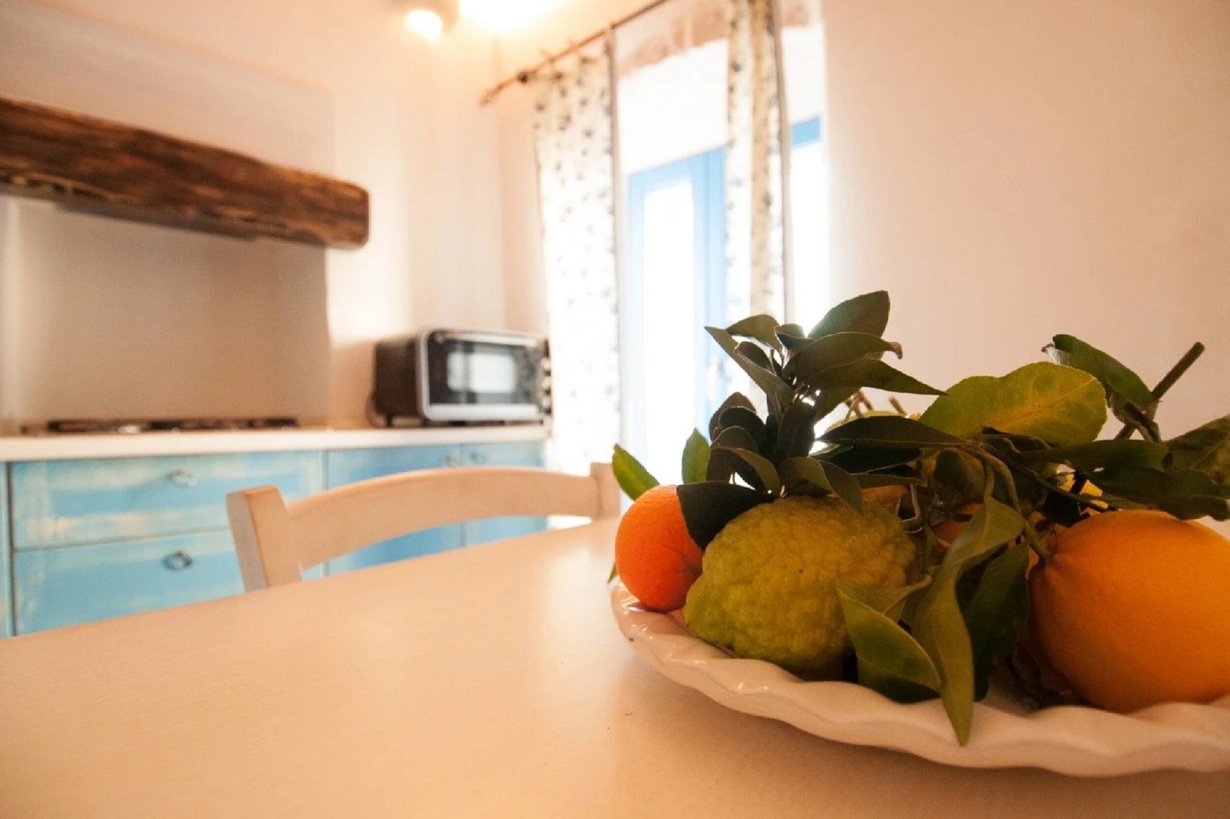 Mesa de jantar com uma fruteira contendo laranjas e uma fruta verde, uma cadeira branca e uma área de cozinha com micro-ondas e armários azuis.
