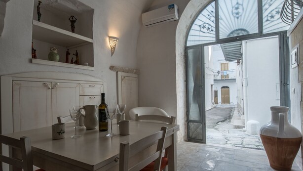 A dining area with a table set for two, a white chair, and a large vase. There is a shelf with decorative items above the table. - Casa Daniele (Ostuni)